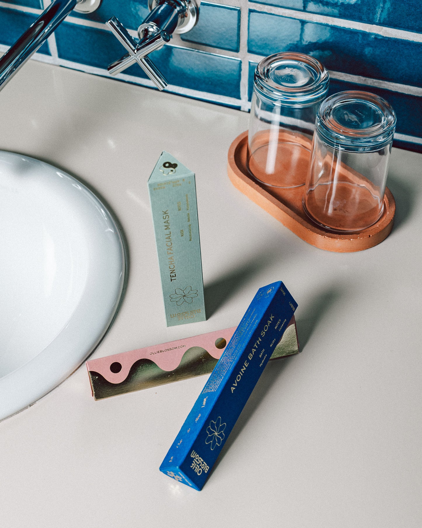 Photograph of boxes of Ollie Blossom products on bathroom counter with part of the sink and knob showing. There are two drinking glasses sitting atop a terracotta holder. Blue tile in the background