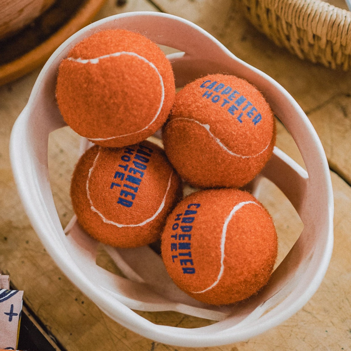 A pile of orange tennis style dog balls with 'CARPENTER HOTEL' printed in blue text on it in a decorative bowl on top of a table.