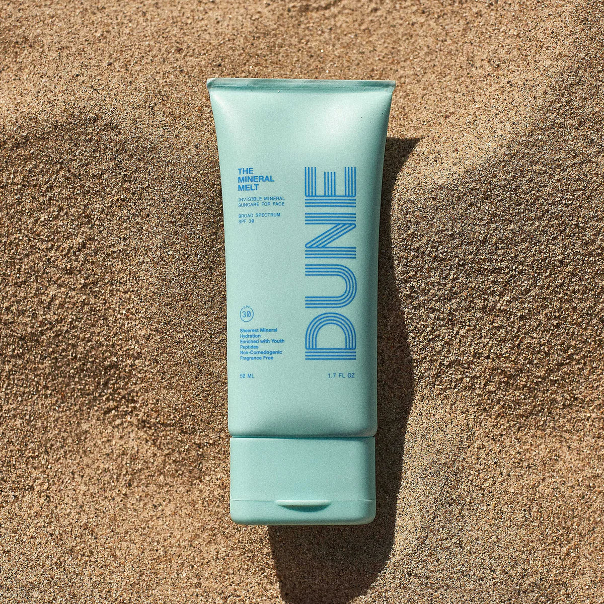 A tube of Dune Suncare The Mineral Melt sunscreen with a light blue color and the brand's logo on the front laying on top of sand