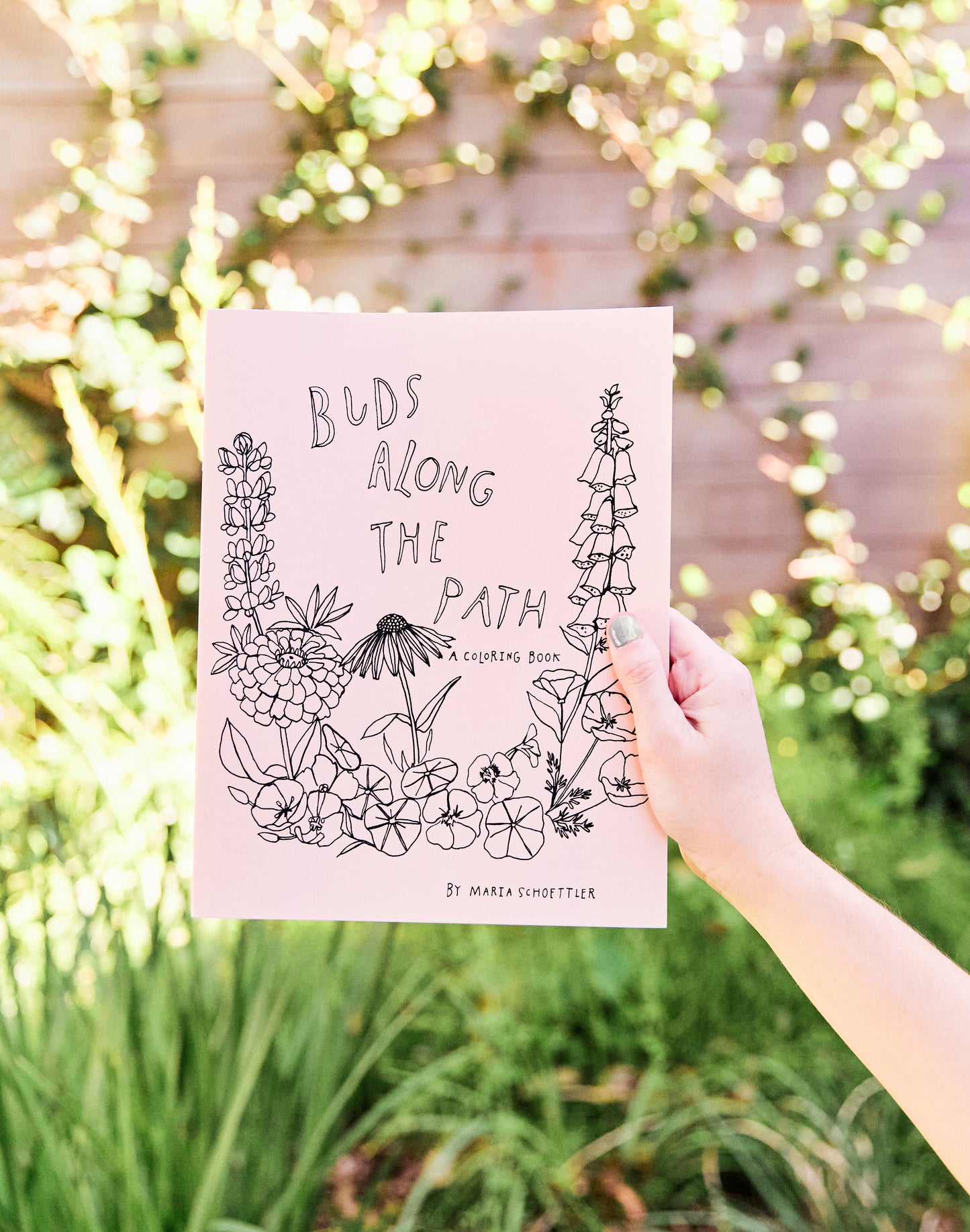 Hand holding a coloring book titled 'Buds Along the Path' against a natural background.