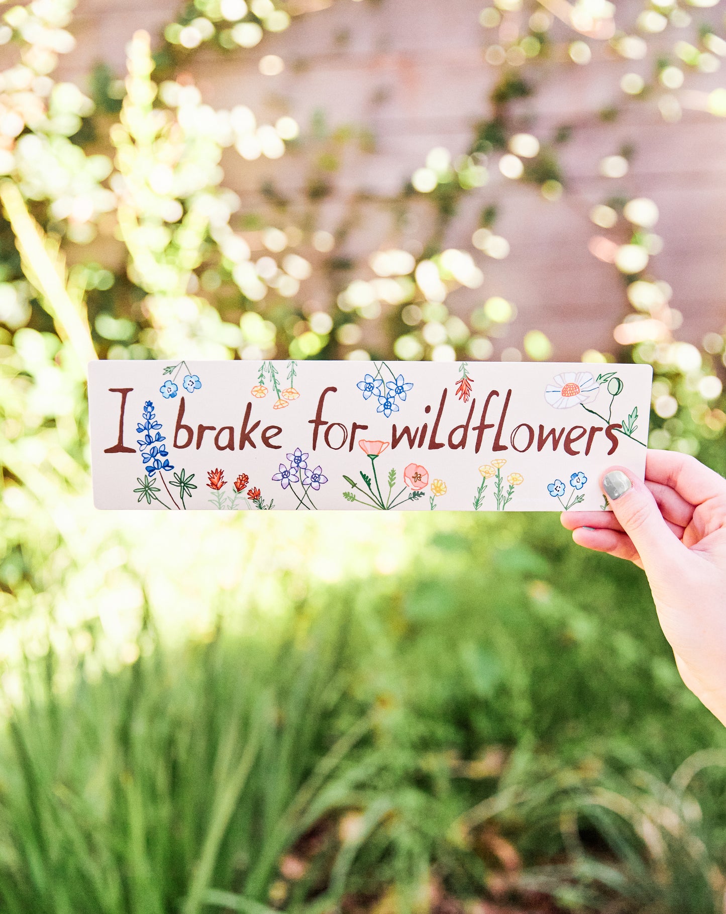 Hand holding a bumpersticker with florals and 'I brake for wildflowers' text against a blurred natural background