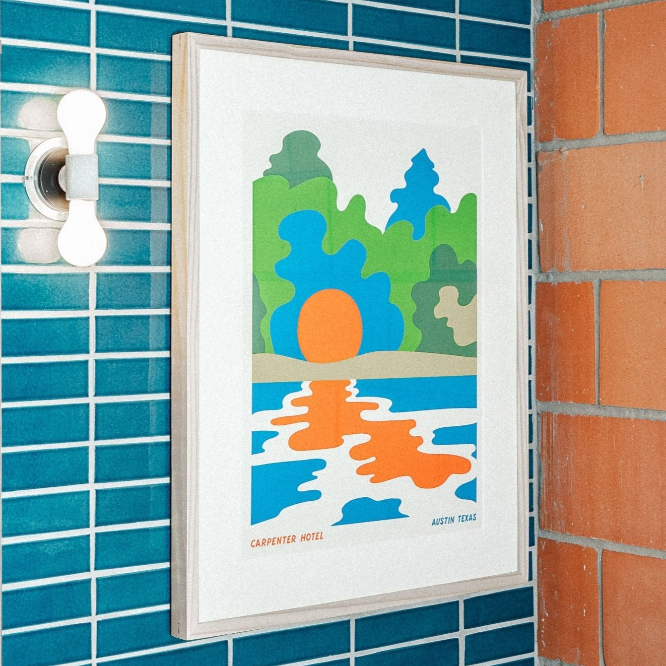 Photograph of graphic poster depicting a lake in front of greenery and the sun hanging in a blue and terracotta tiled hotel bathroom at carpenter hotel.
