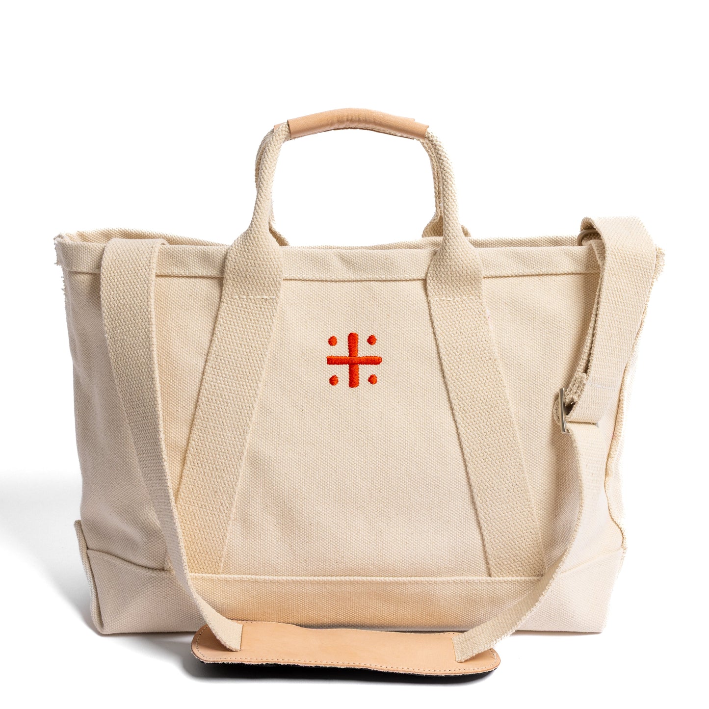 A beige canvas tote bag with leather handles and a red cross logo on the front with shoulder strap featured in the front