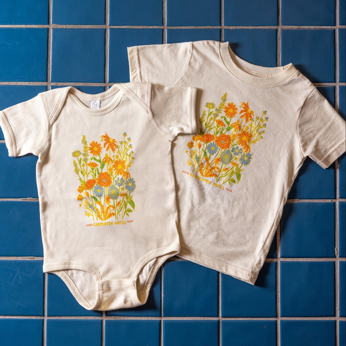 A beige baby onesie and kids t-shirt with a colorful floral pattern and the text 'Carpenter Hotel austin texas' printed on it laying on top of blue tile