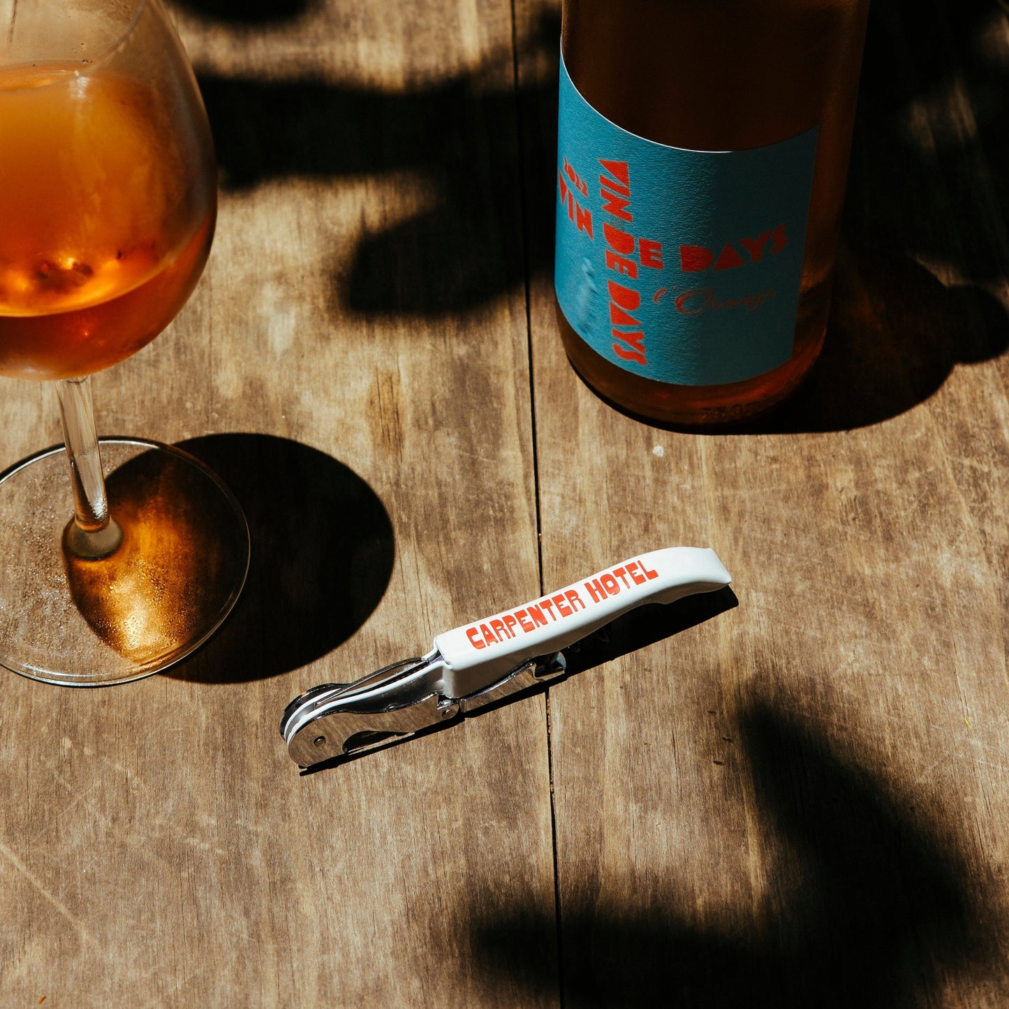 A silver metal wine key with the Carpenter Hotel logo printed in red on its side sitting on a wooden table alongside a glass and bottle of wine