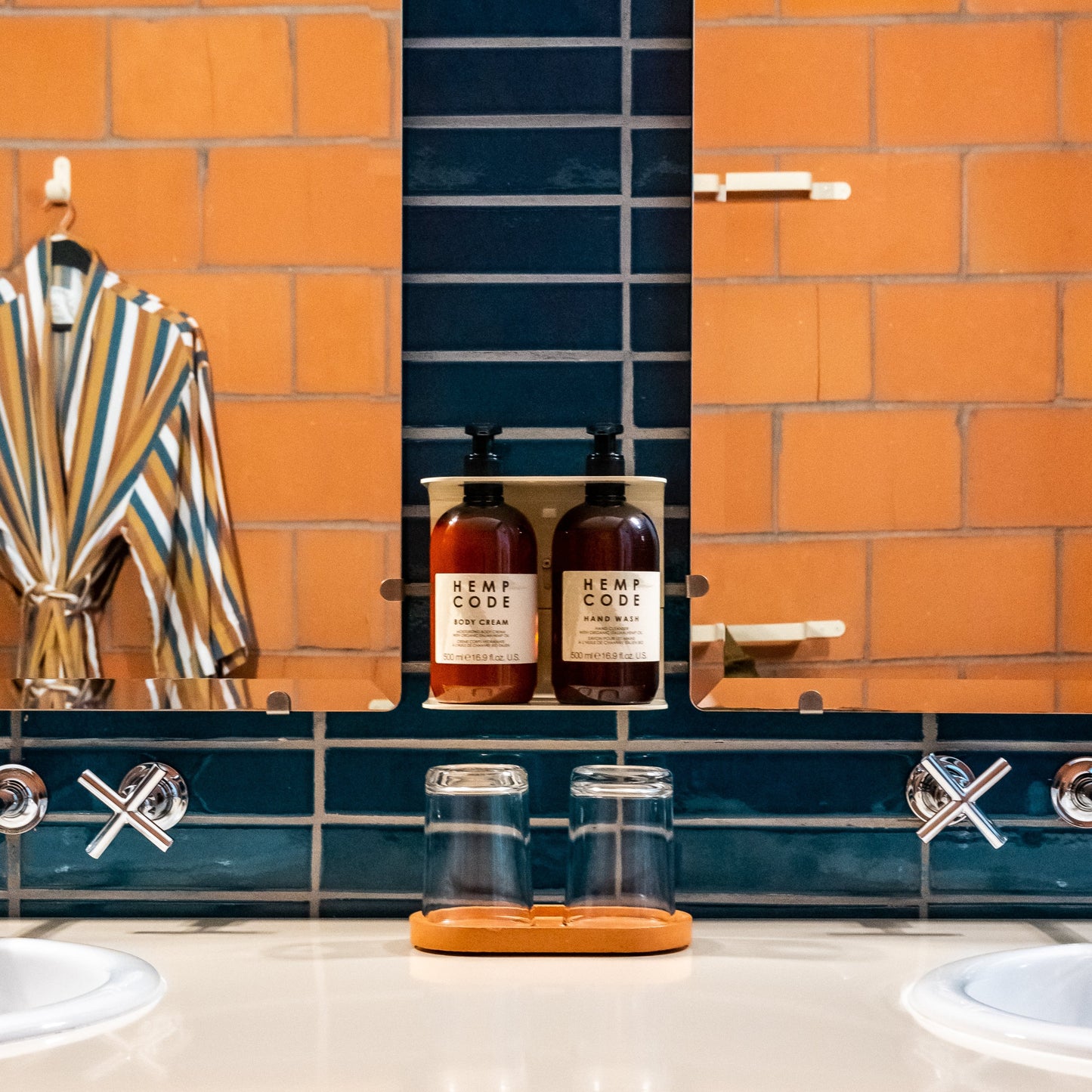 Photograph of Hemp Code hand wash and body cream in wall stand in a hotel bathroom with blue tiles. There is a mirror on either side reflecting the orange tiles and bathrobe to create an illusion