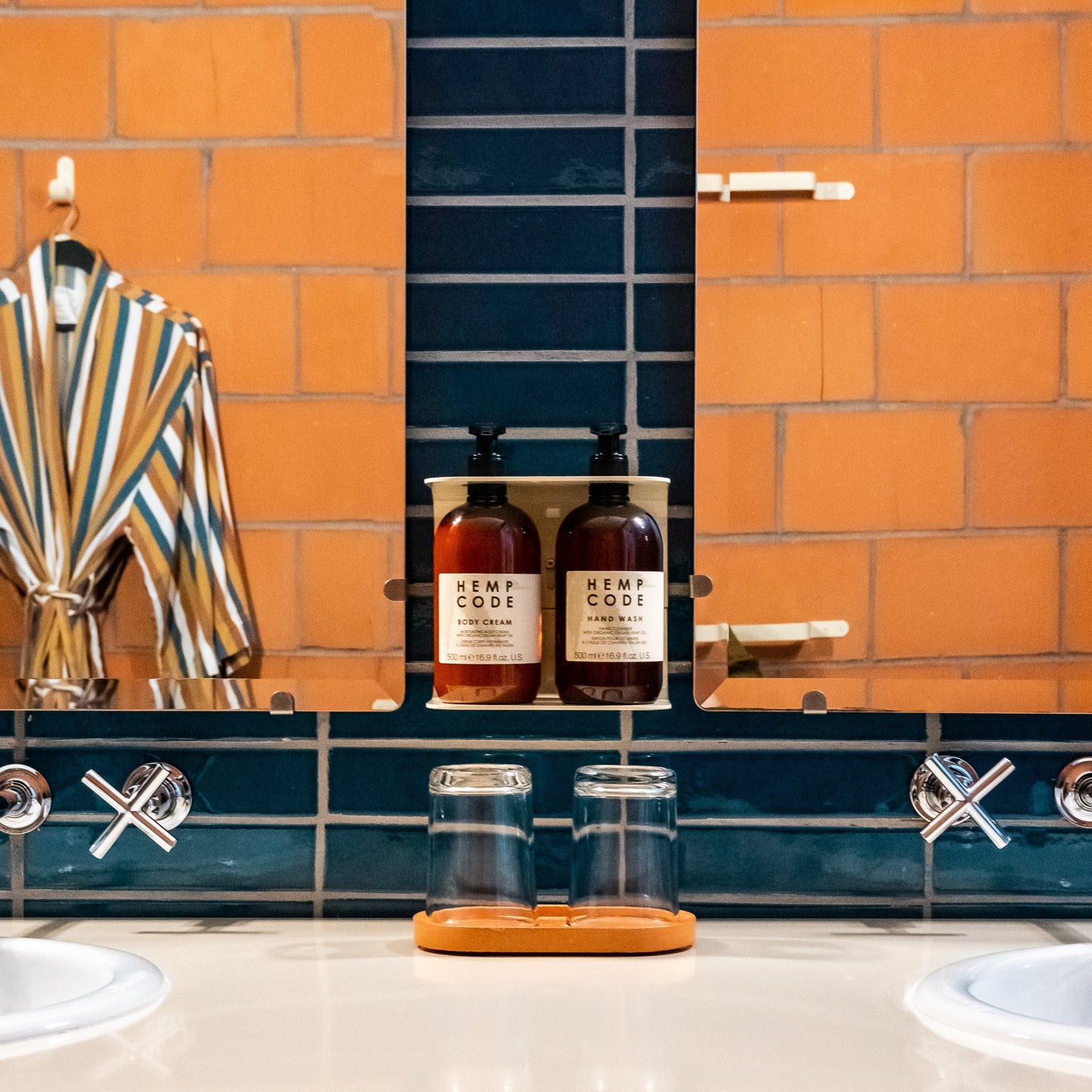 Photograph of Hemp Code hand wash and body cream in wall stand in a hotel bathroom with blue tiles. There is a mirror on either side reflecting the orange tiles and bathrobe to create an illusion