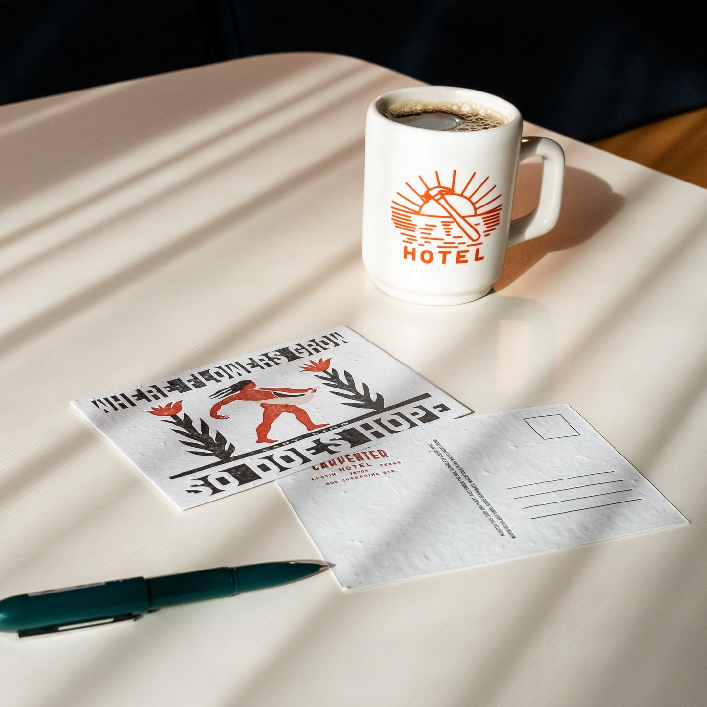A postcard with letterpress text and illustration, featuring a figure sowing seeds among flowers, with the text 'WHERE FLOWERS GROW SO DOES HOPE - LADY BIRD' pictured with a pen and mug of coffee on top of beige table.