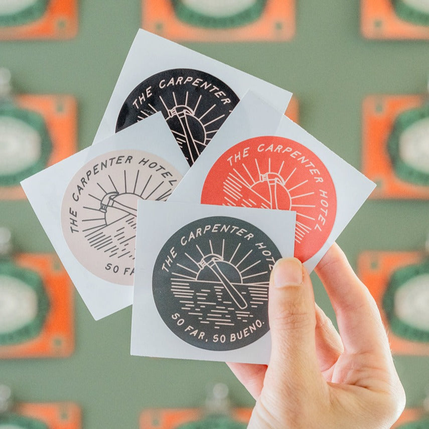 A hand holding four round stickers with a sun motif and the text 'THE CARPENTER HOTEL SO FAR SO BUENO'. The stickers come in different colors including red, olive, black, and tan.