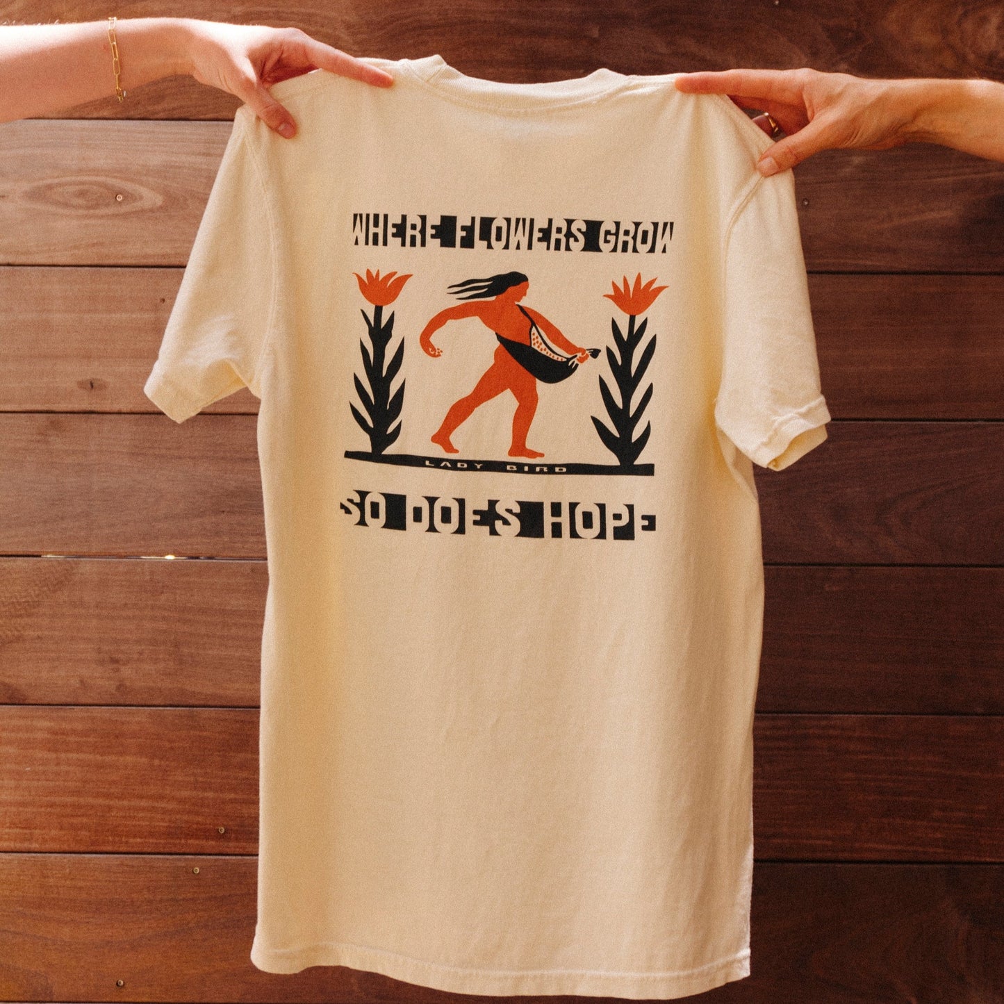 Beige tshirt with black and orange graphic of person carrying seeds with tall flower plant on either side. Text reads 'WHERE FLOWERS GROW SO DOES HOPE' being held by two hands against a wooden background