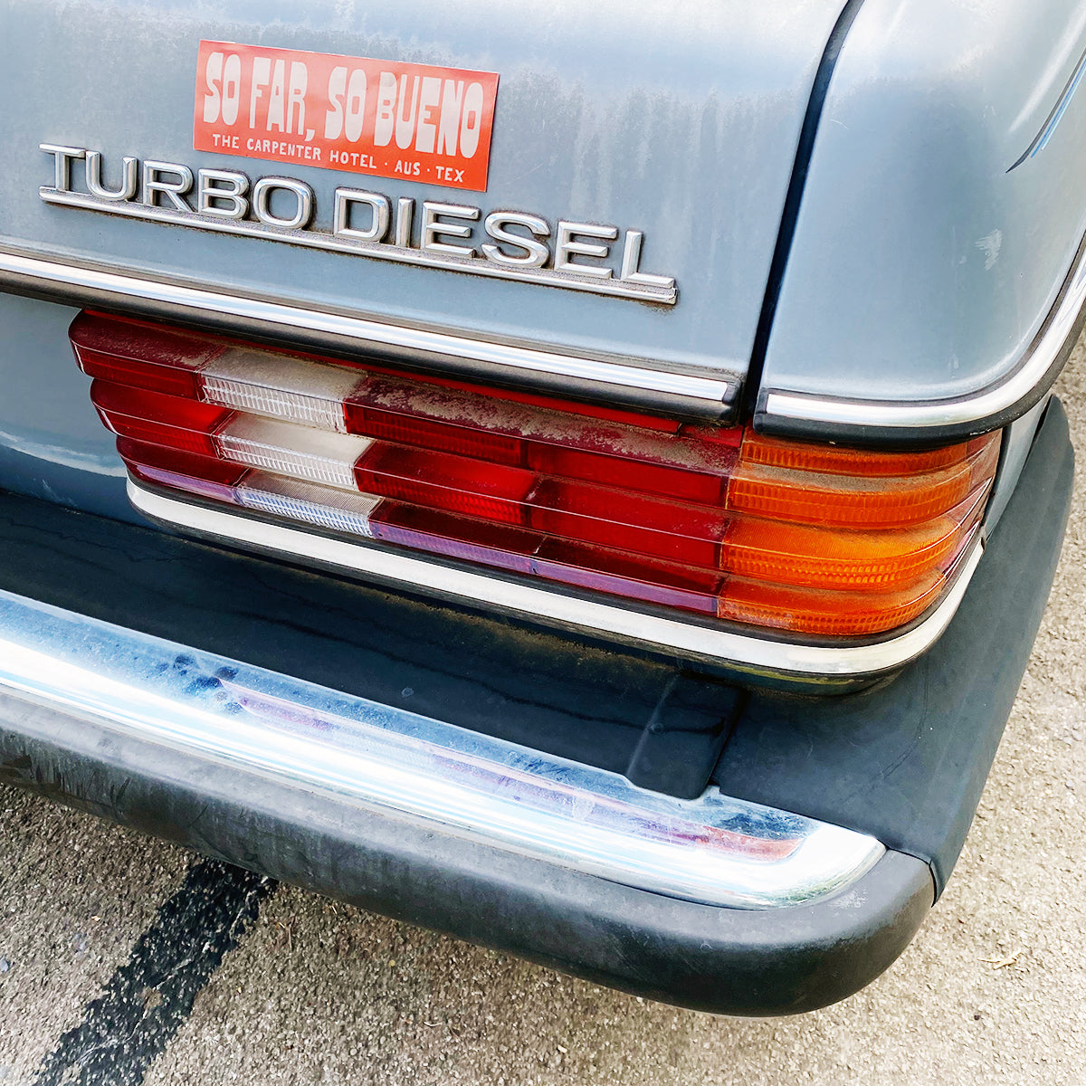 An orange and beige bumper sticker with the text 'SO FAR, SO BUENO THE CARPENTER HOTEL AUST TEX' on the back of light blue vintage car.