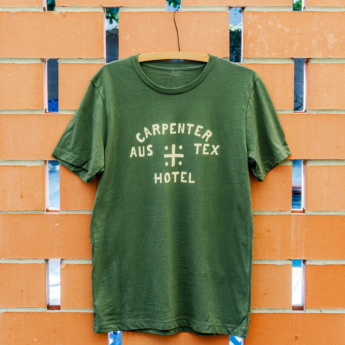 Green tshirt with 'CARPENTER HOTEL AUS TEX' on front in beige graphic. The shirt is hanging on wooden hanger against a terracotta brick background.
