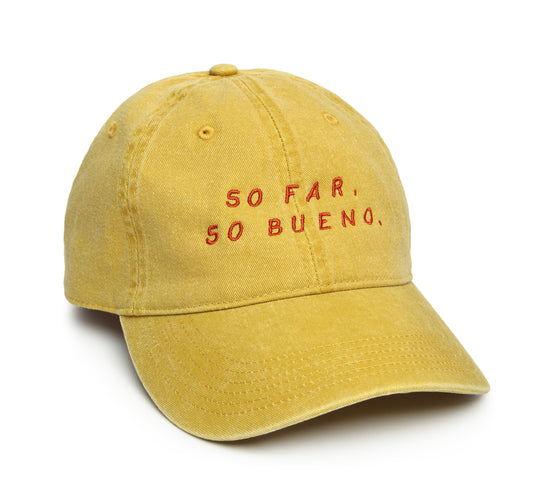 A yellow 'dad hat' with red embroidered text that reads 'SO FAR, SO BUENO'.
