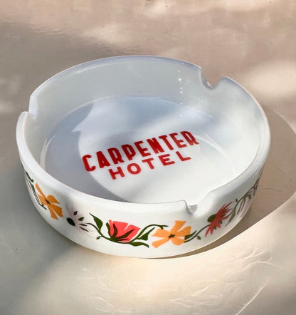 White ceramic ashtray with floral designs  on outside and 'CARPENTER HOTEL' text in center bottom sitting on a light background