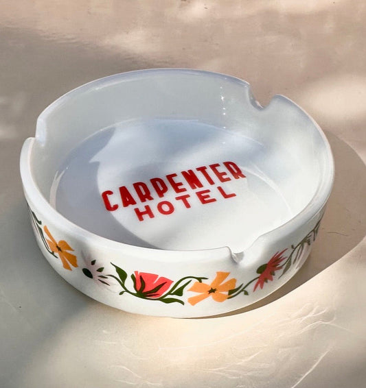 White ceramic ashtray with floral designs  on outside and 'CARPENTER HOTEL' text in center bottom sitting on a light background