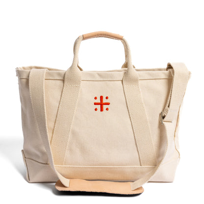 A beige canvas tote bag with leather handles and a red cross logo on the front with shoulder strap featured in the front 