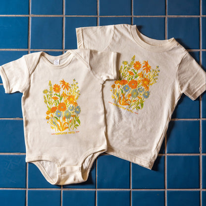 A beige baby onesie and kids t-shirt with a colorful floral pattern and the text 'Carpenter Hotel austin texas' printed on it laying on top of blue tile