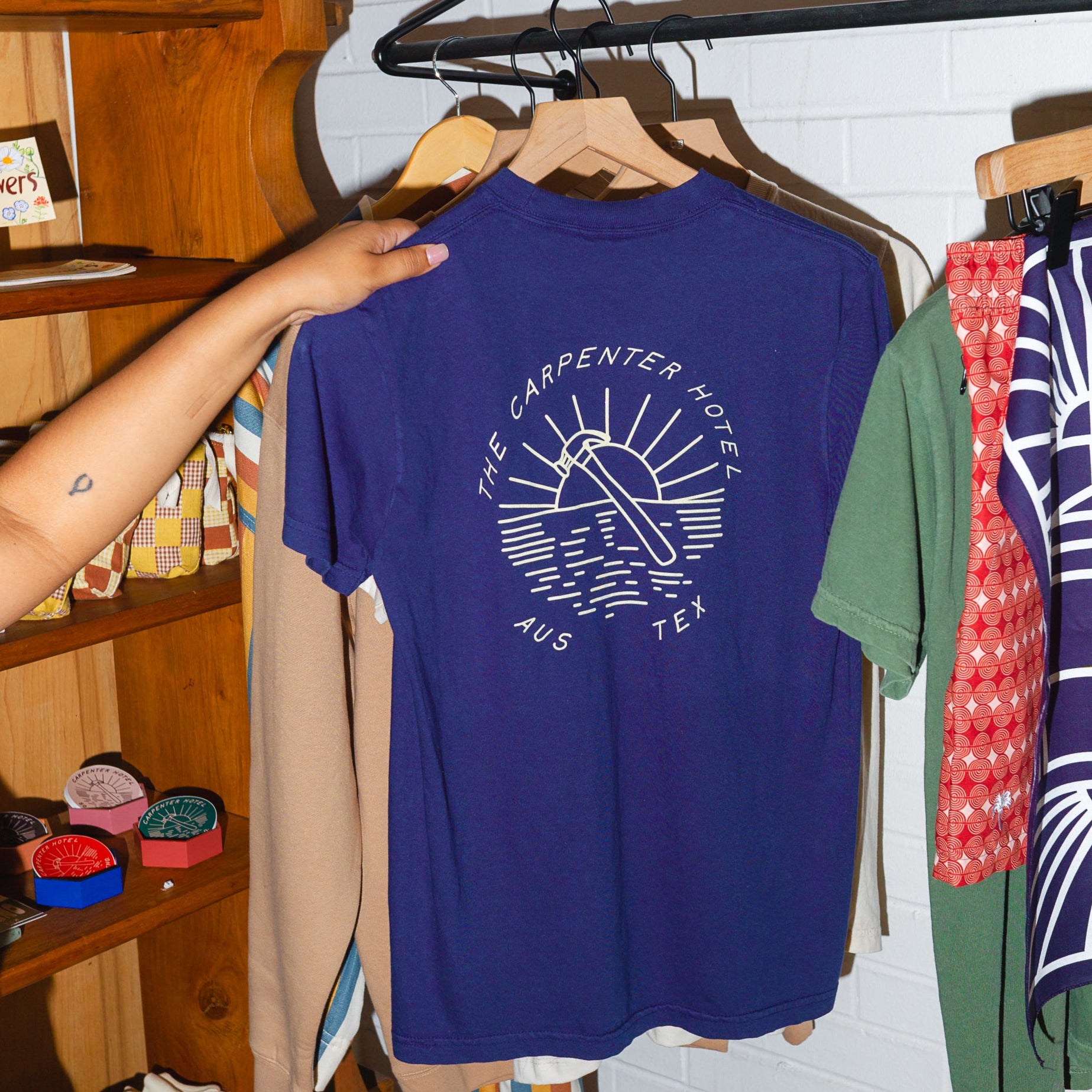 Photograph of navy blue tshirt that says 'THE CARPENTER HOTEL AUS TEX' with line drawing of hammer and sunset in center. The shirt is hanging on a wooden hanger, being held by someone with wooden shelves with other products in the background.