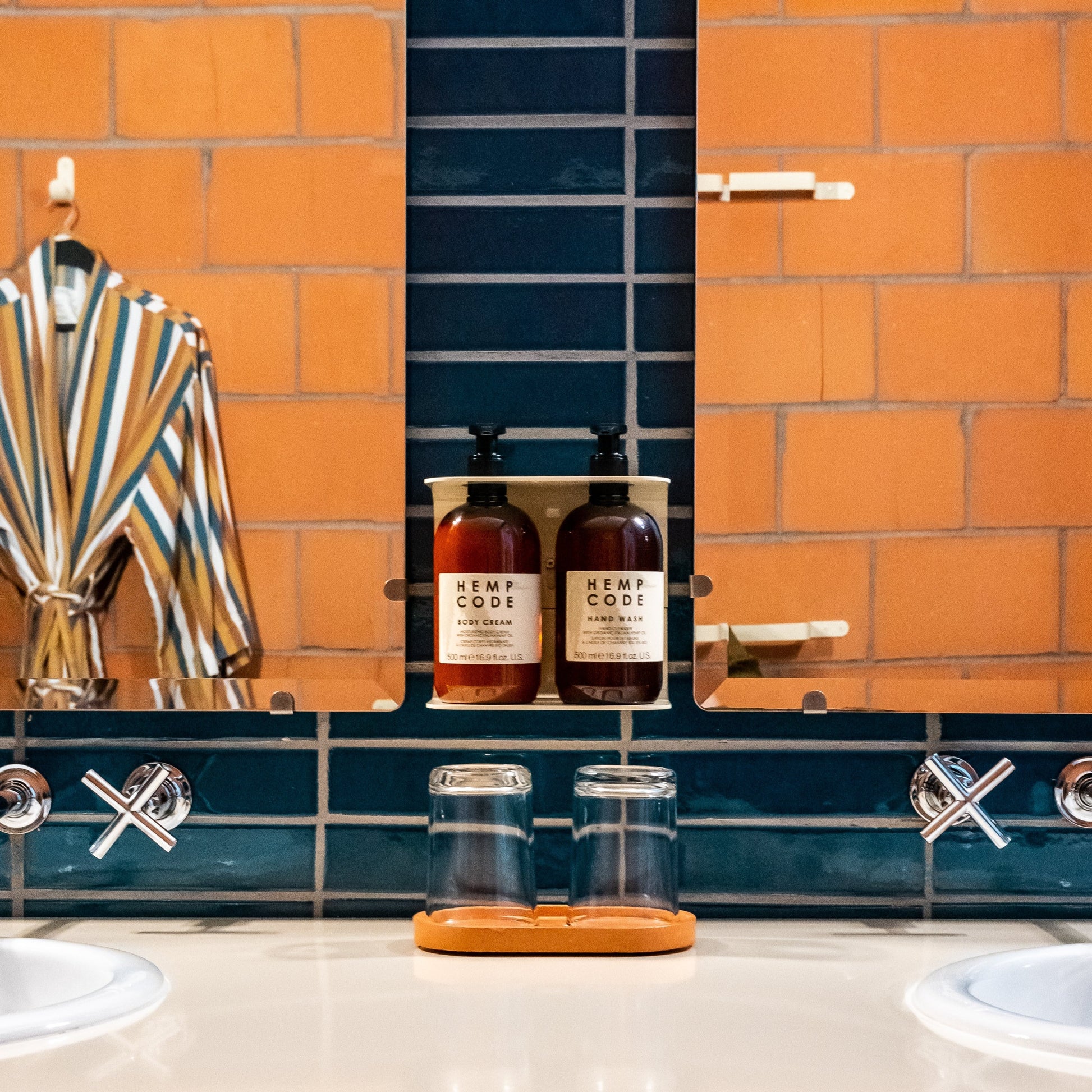 Photograph of Hemp Code hand wash and body cream in wall stand in a hotel bathroom with blue tiles. There is a mirror on either side reflecting the orange tiles and bathrobe to create an illusion