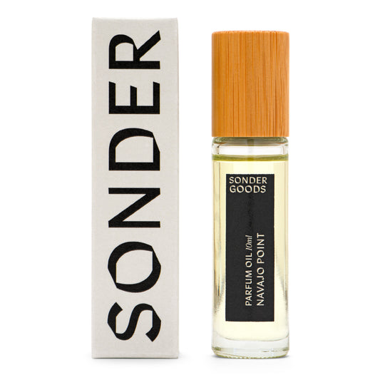 A small, clear glass bottle with a wooden cap, labeled 'Sonder Goods Parfum Oil Navajo Point', next to its white box packaging with the word 'SONDER' in black text.