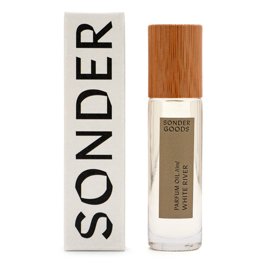 A small, clear glass bottle with a wooden cap, labeled 'Sonder Goods Parfum Oil White River', next to its white box packaging with the word 'SONDER' in black text.
