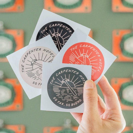 A hand holding four round stickers with a sun motif and the text 'THE CARPENTER HOTEL SO FAR SO BUENO'. The stickers come in different colors including red, olive, black, and tan.